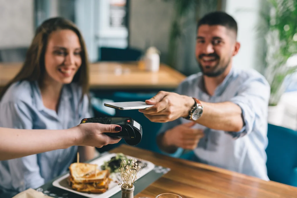 Man using a smartphone to make a contactless crypto payment at a restaurant while seated with a woman, both smiling and enjoying their dining experience.
