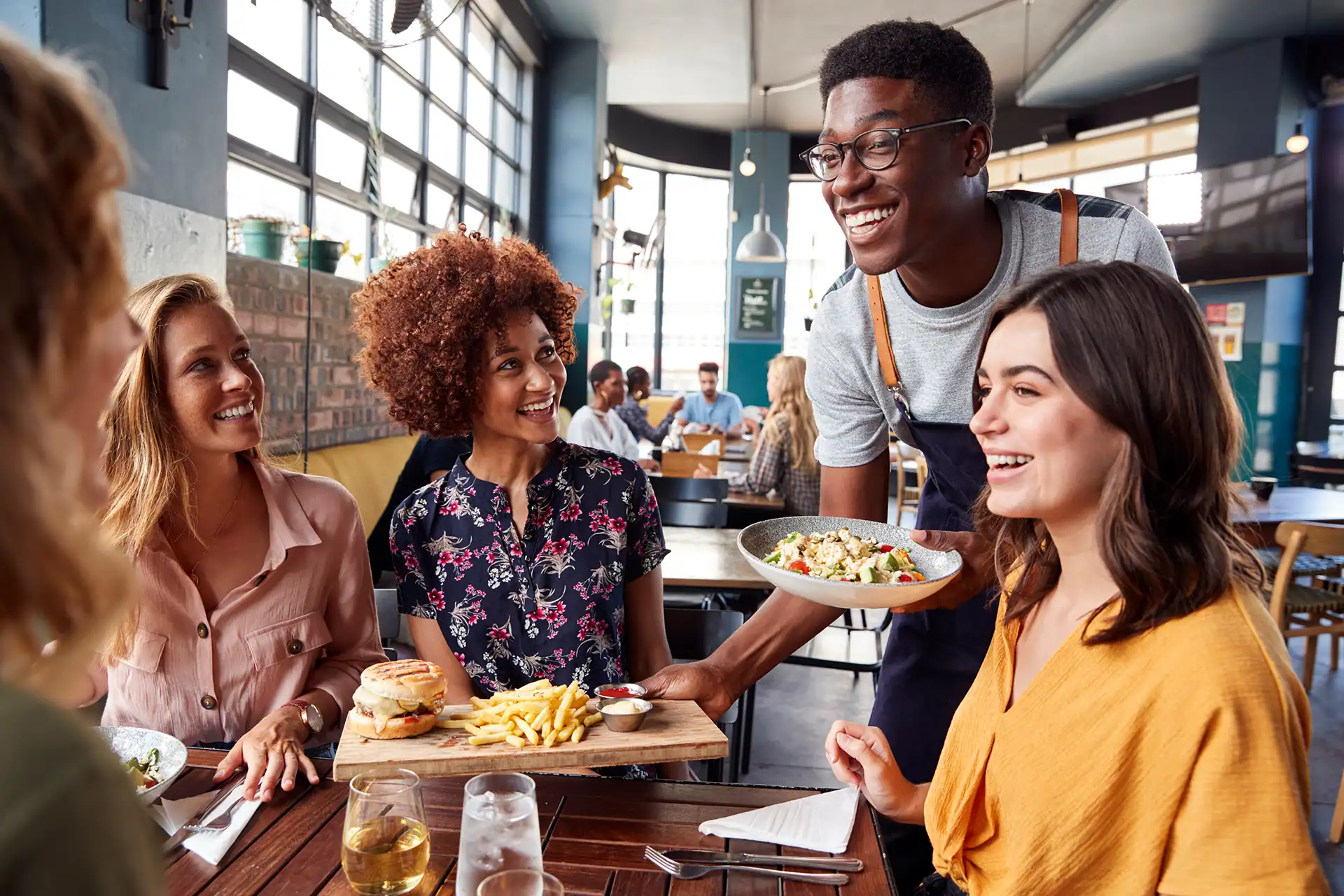 A smiling waiter serves a cheerful group of young women at a modern, casual restaurant during the daytime, bringing value to the group.