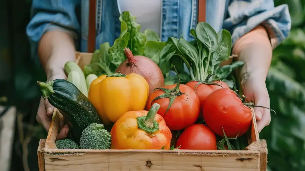 Person holding a wooden crate filled with fresh, locally grown vegetables including tomatoes, bell peppers, zucchini, broccoli, and leafy greens to emphasize sustainability.